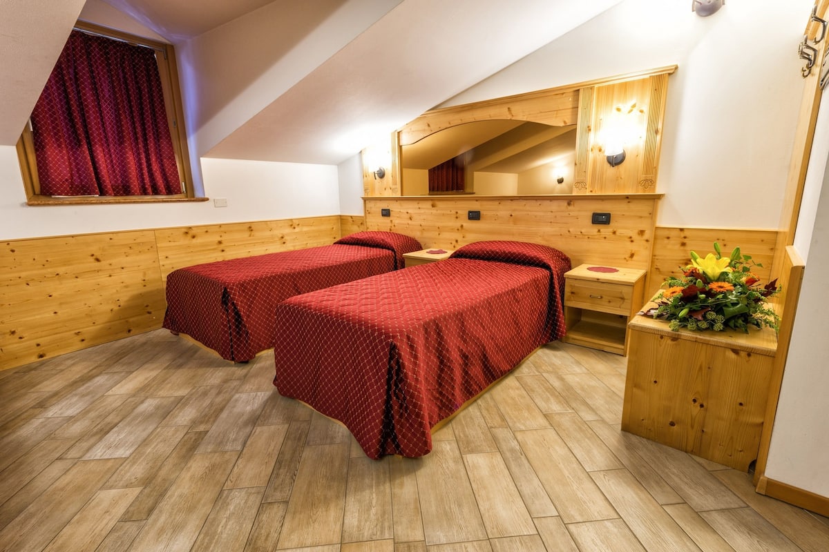 A room with two beds, wooden walls, a wooden floor, a window with red curtains, a mirror, and a small table with a plant.