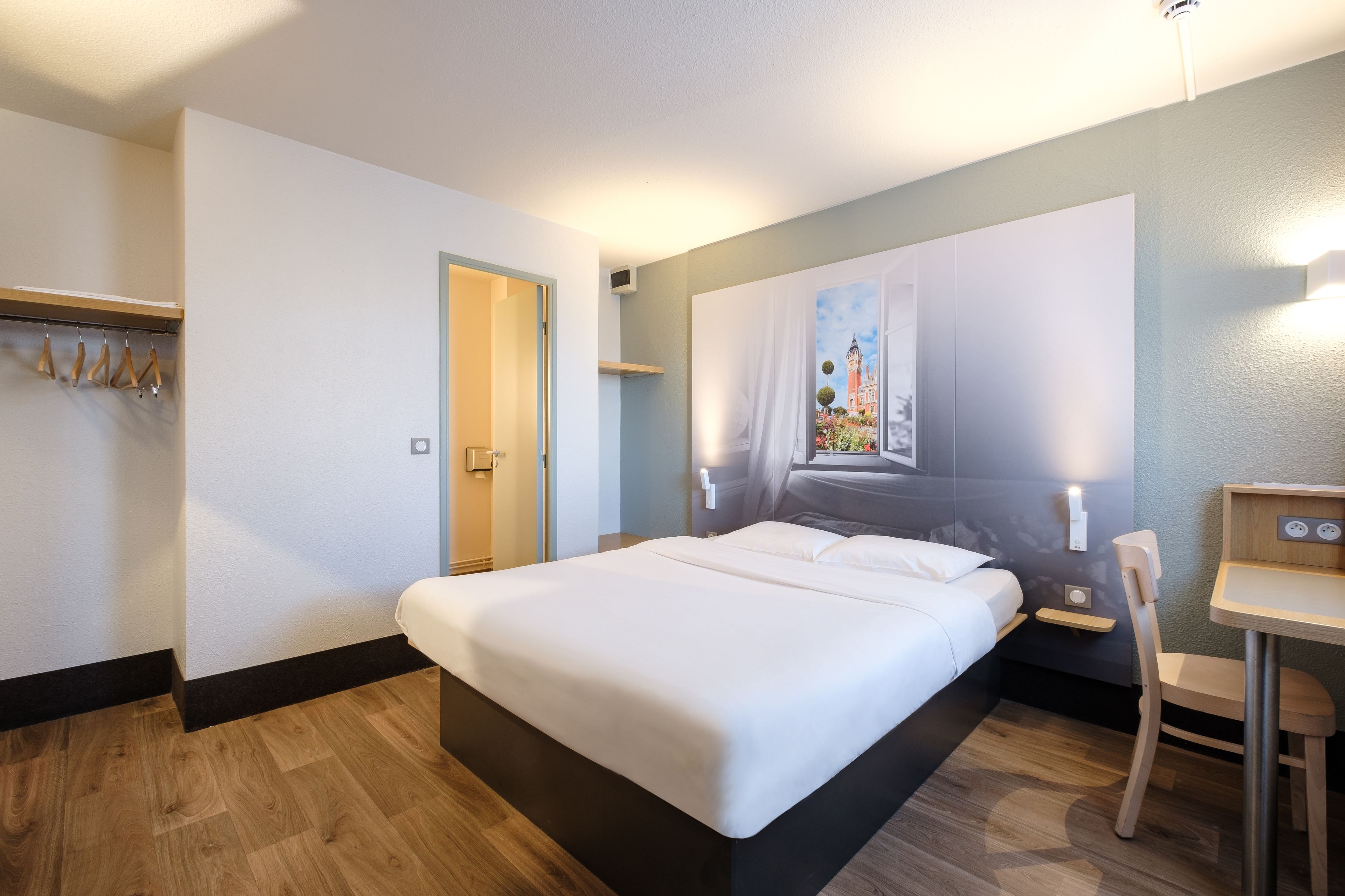 Habitación de hotel con una cama grande, un escritorio y una silla. Se observa un perchero con perchas y una puerta que da a otra habitación.
