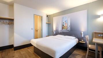 Une chambre d’hôtel avec un grand lit, un bureau et une chaise. On y trouve un porte-manteau avec des cintres et une porte qui donne sur une autre pièce.