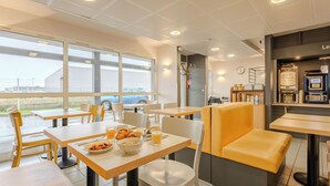 A modern cafe with wooden tables and chairs, a seating area with yellow cushions, and a view of the outside through large windows. - B&B HOTEL Calais Coquelles Tunnel sous la Manche (Coquelles)