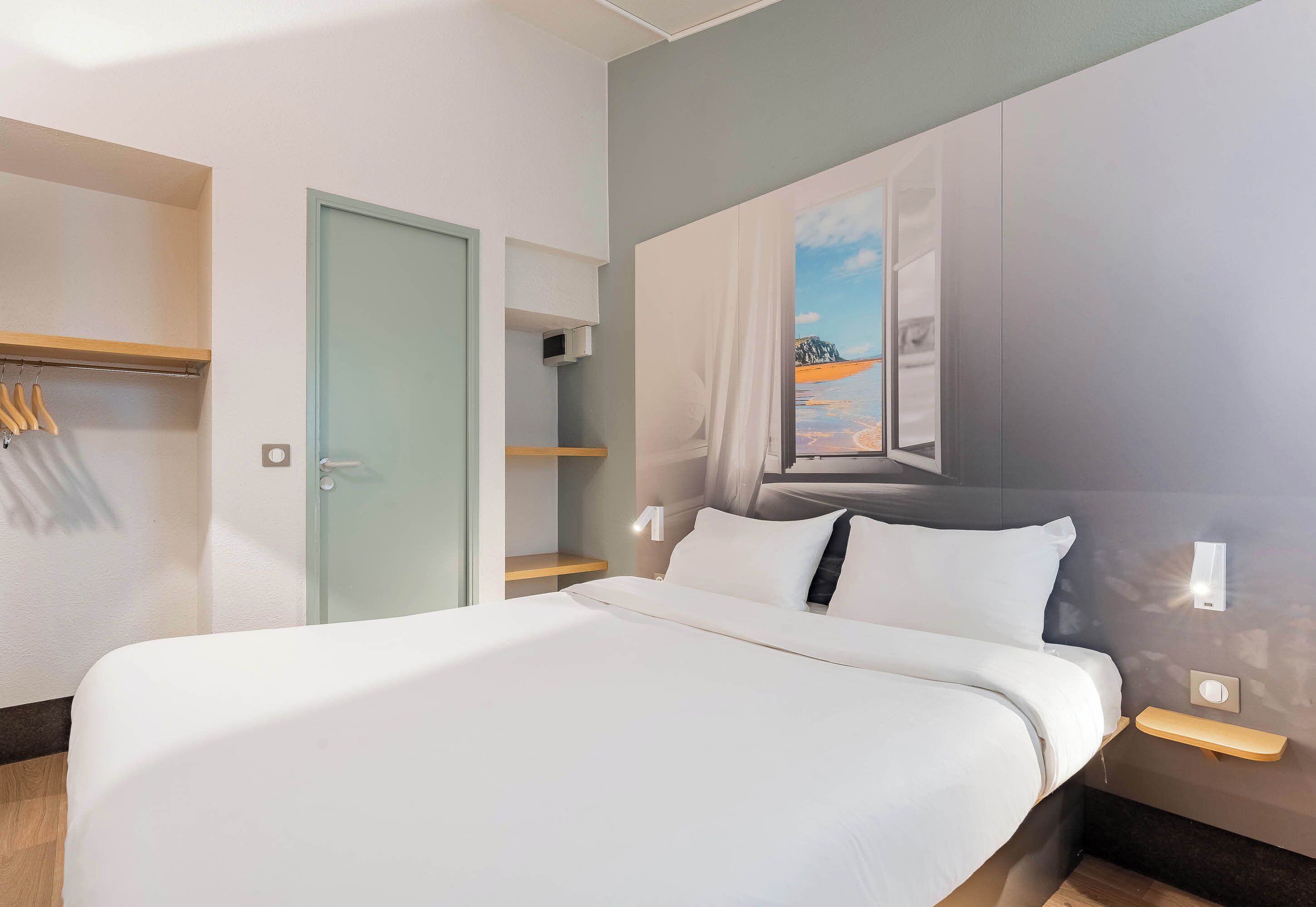 Une chambre d’hôtel moderne avec un grand lit, une armoire, un miroir et un tableau représentant une scène de plage.