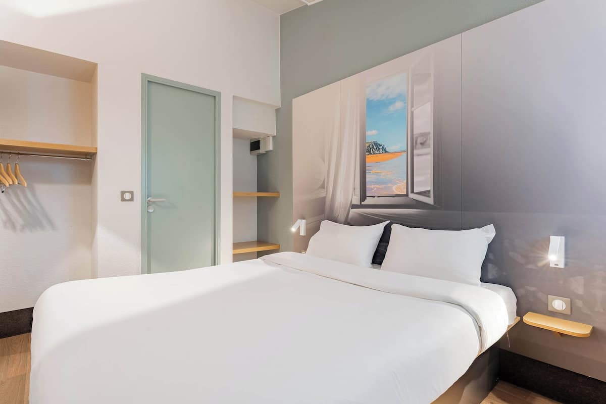 Une chambre d’hôtel moderne avec un grand lit, une armoire, un miroir et un tableau représentant une scène de plage.