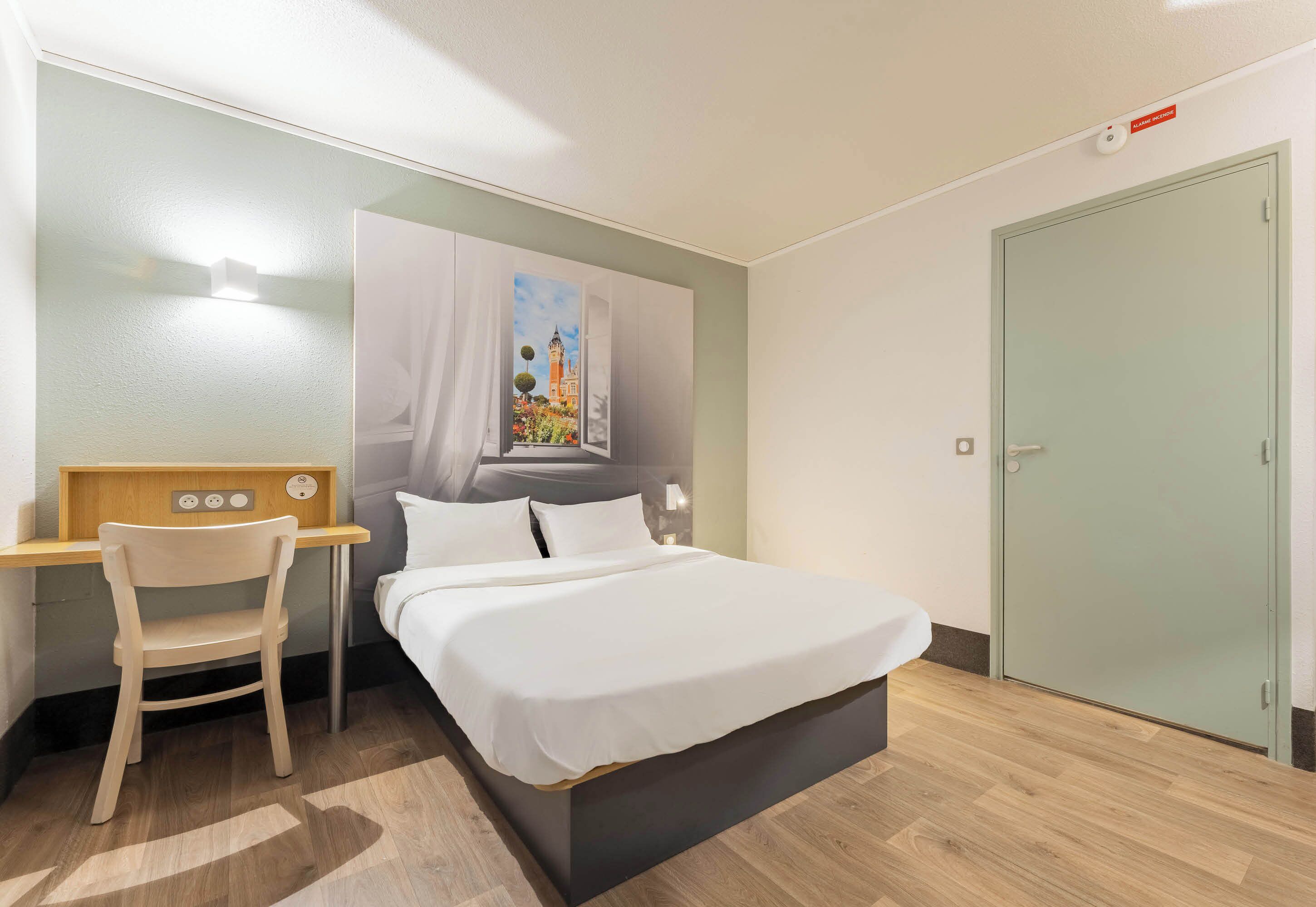 A hotel room with a bed, a desk with a chair, a painting on the wall, and a door.