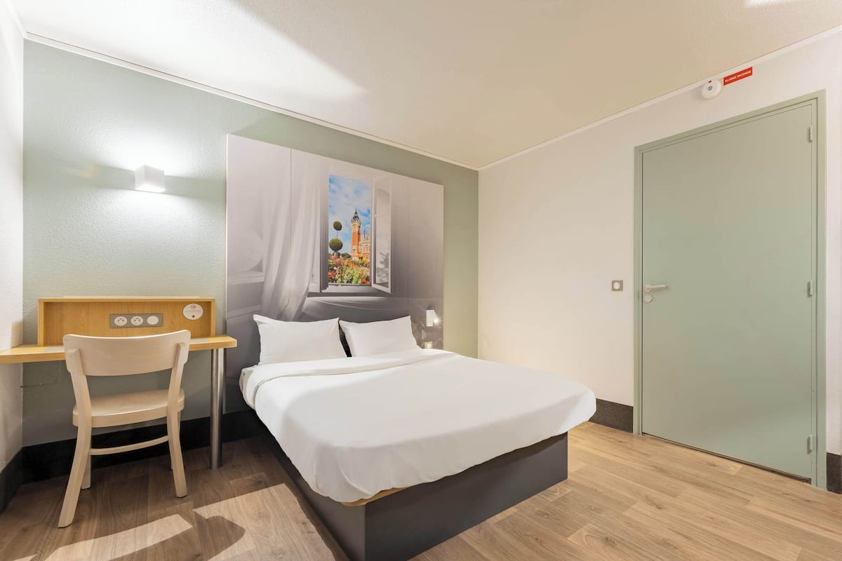 A hotel room with a bed, a desk with a chair, a painting on the wall, and a door.