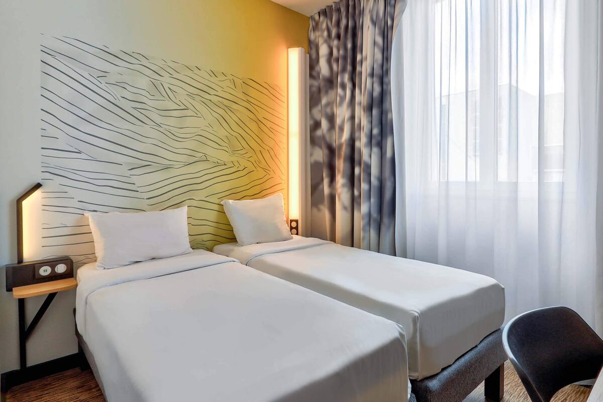 A hotel room with two beds, a nightstand with a radio, and a large window with sheer curtains.
