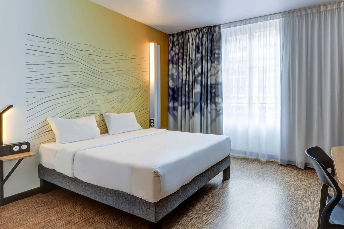 A modern hotel room with a large bed, a bedside table with a lamp, a wall art piece, and a view of the outdoors through sheer curtains.