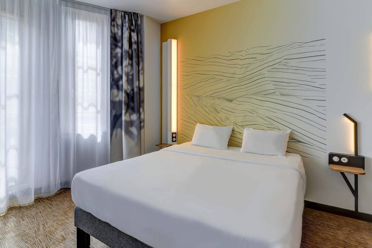 A hotel room with a large bed, white bedding, a modern wall art piece, a bedside table with a lamp, and sheer curtains on the window.
