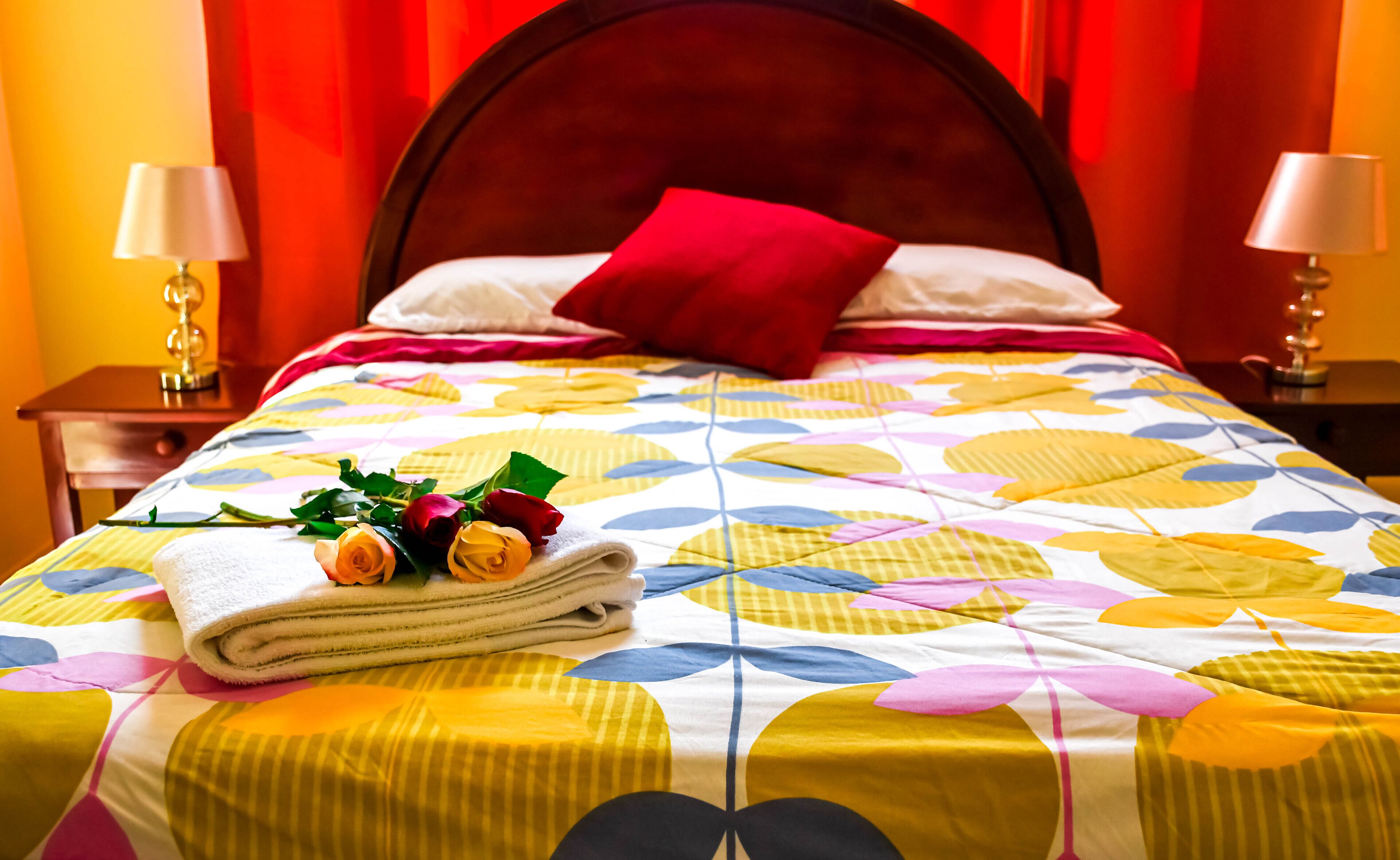 A neatly made bed with a colorful floral bedspread, a red pillow, and a stack of towels with roses.