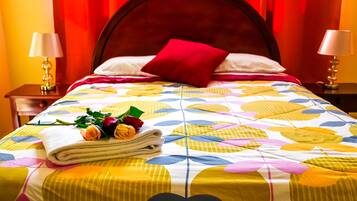 A neatly made bed with a colorful floral bedspread, a red pillow, and a stack of towels with roses.
