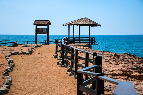 A beach path leading to a wooden gazebo with a signboard, surrounded by a wooden fence and a stone wall. - Hotel Suites Costa de Oro (La Libertad)