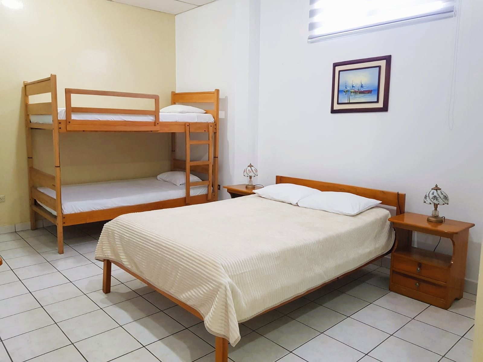 A bedroom with a bunk bed, a single bed, a nightstand with a lamp, and a framed picture on the wall.