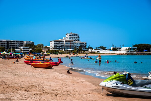 Una playa con kayaks y un jet ski, una orilla arenosa y edificios al fondo.