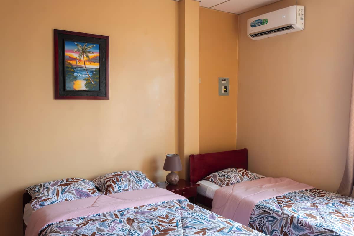 Camera d'albergo con due letti, un quadro raffigurante un tramonto, un condizionatore d'aria e un comodino con una lampada.