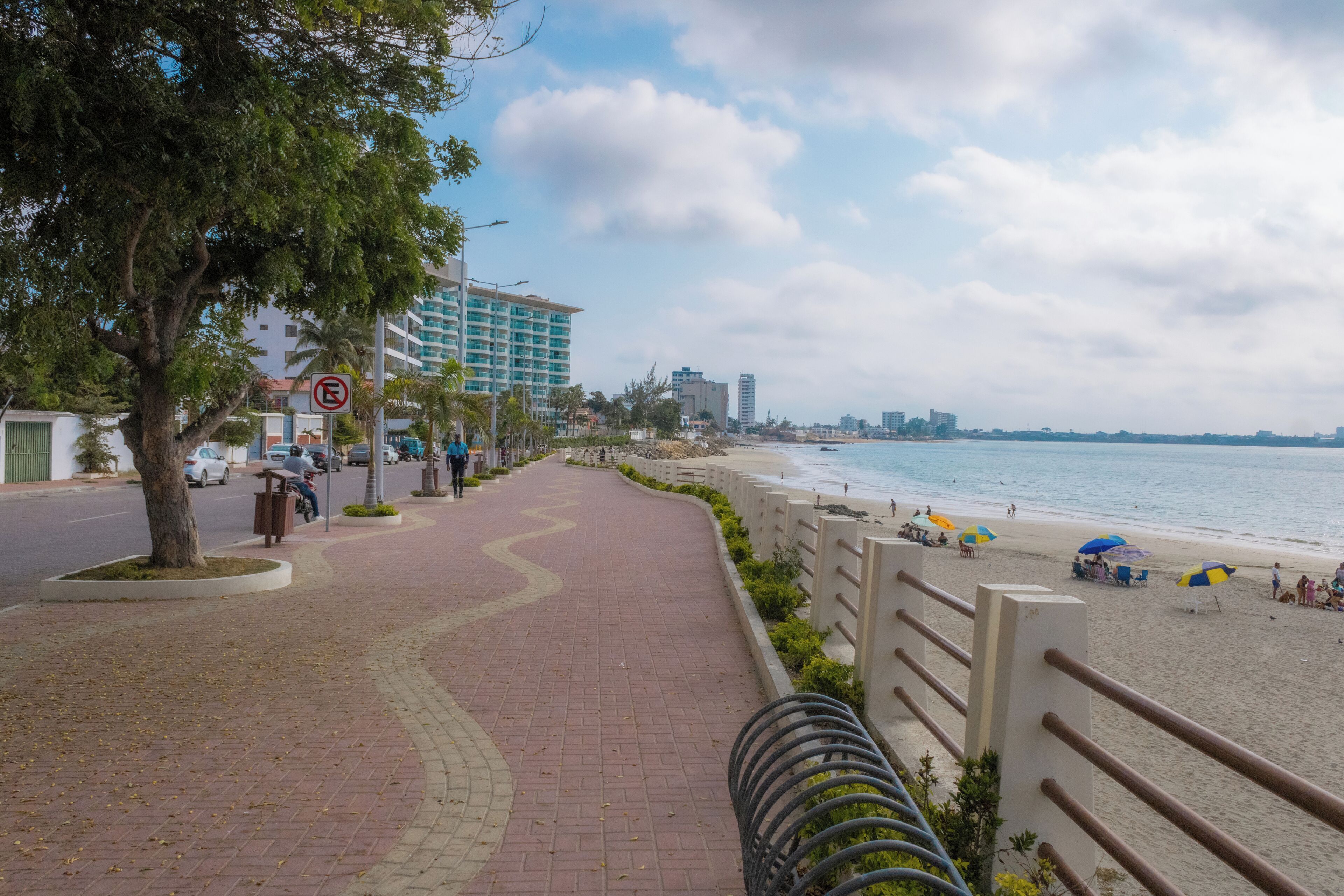 A coastal walkway with a beach, palm trees, and a hotel in the background.
