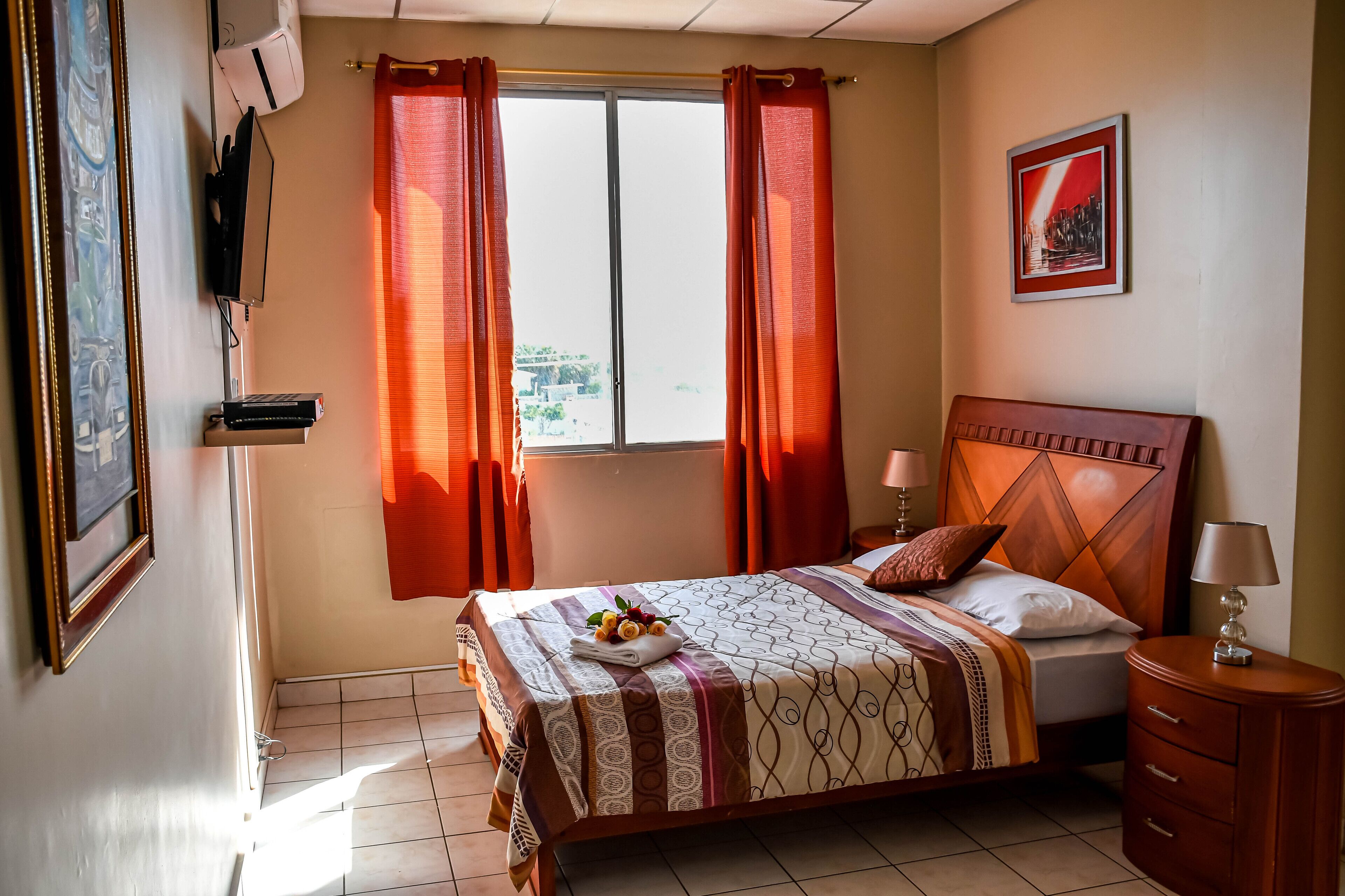 Une chambre à coucher bien rangée, avec un lit, une table de chevet, une lampe et une fenêtre avec des rideaux.