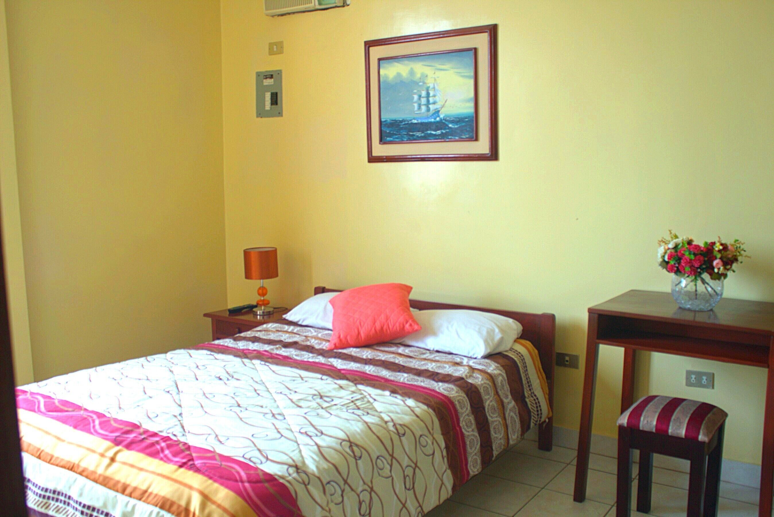 A neatly made bed with a patterned bedspread and pillows, a bedside table with a lamp, a framed picture on the wall, and a small table with a vase of flowers.