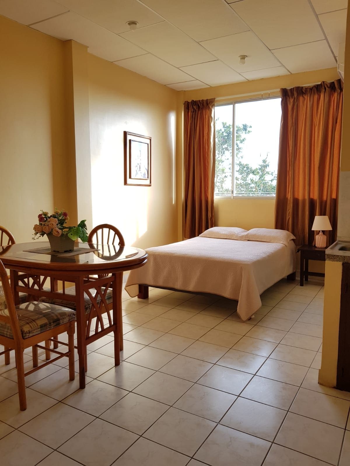 Una habitación de hotel con cama, una mesa de comedor con sillas, una mesita con lámpara y una ventana con cortinas.
