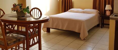 Una habitación de hotel con cama, una mesa de comedor con sillas, una mesita con lámpara y una ventana con cortinas.