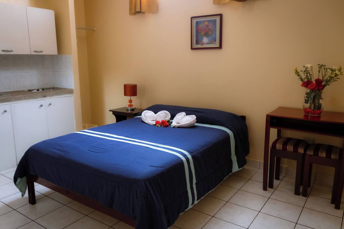 Una camera d'albergo con un letto, un tavolino, una sedia, un comodino, una lampada, un quadro e un vaso con fiori.