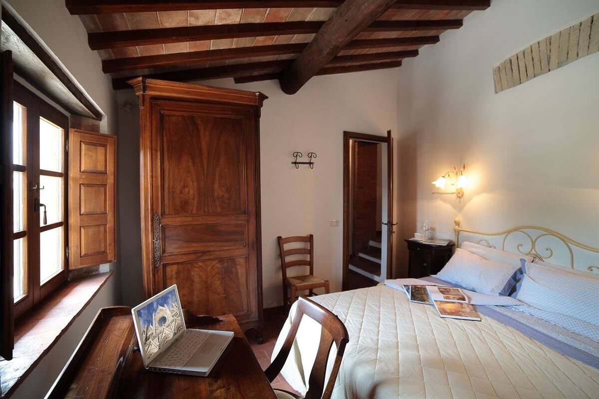 A bedroom with a wooden desk, a bed with a quilt, a chair, a wooden wardrobe, and a wall-mounted light fixture.