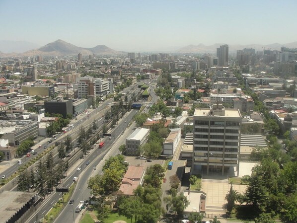 An aerial view of a cityscape with a mix of modern and older buildings, a wide road with vehicles, and a mountain in the background. - Apartment Las Heras (Santiago)