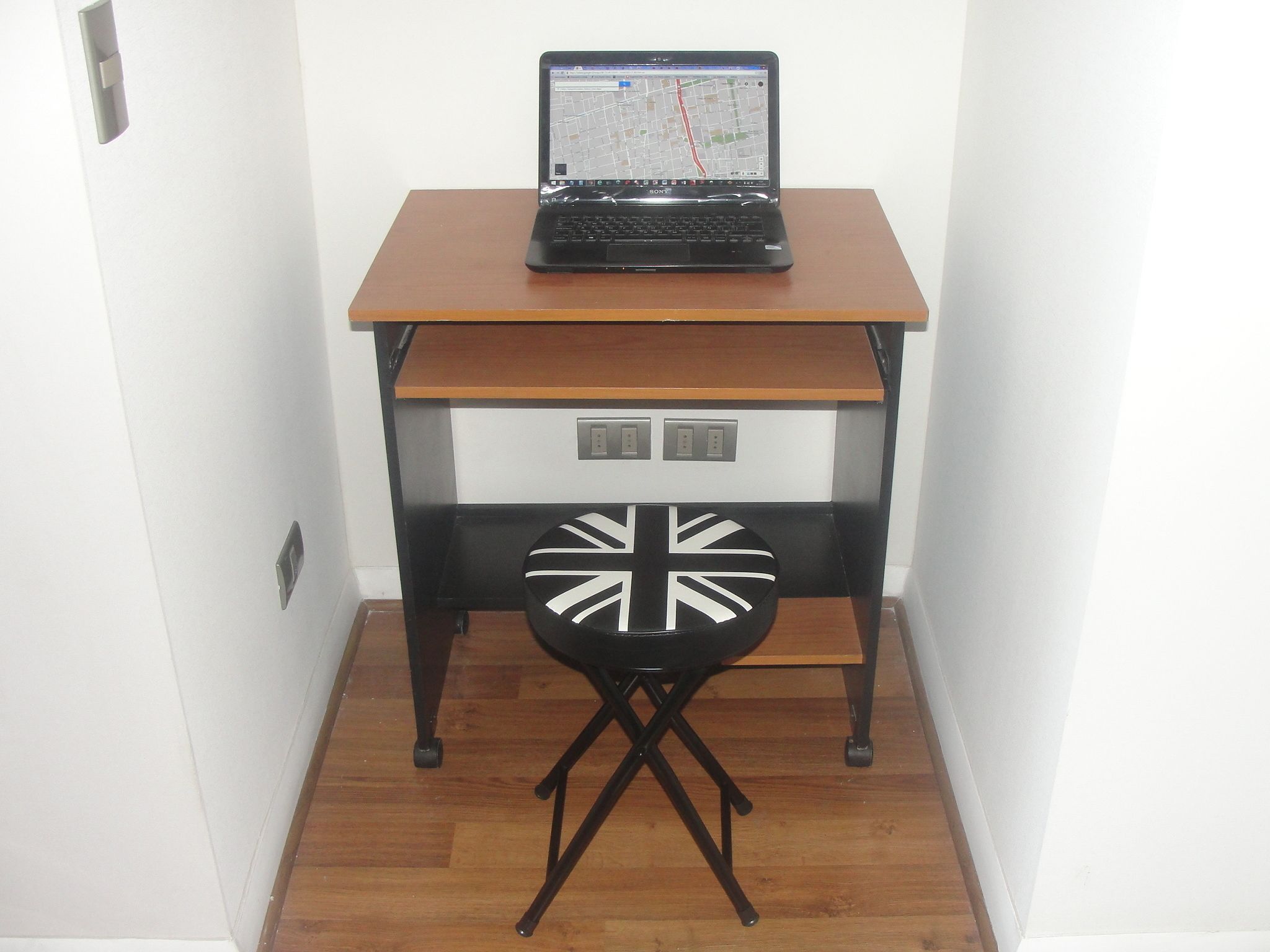 Een laptop op een houten bureau, met een kaart op het scherm, een zwarte stoel met een Union Jack ontwerp en een witte muur.
