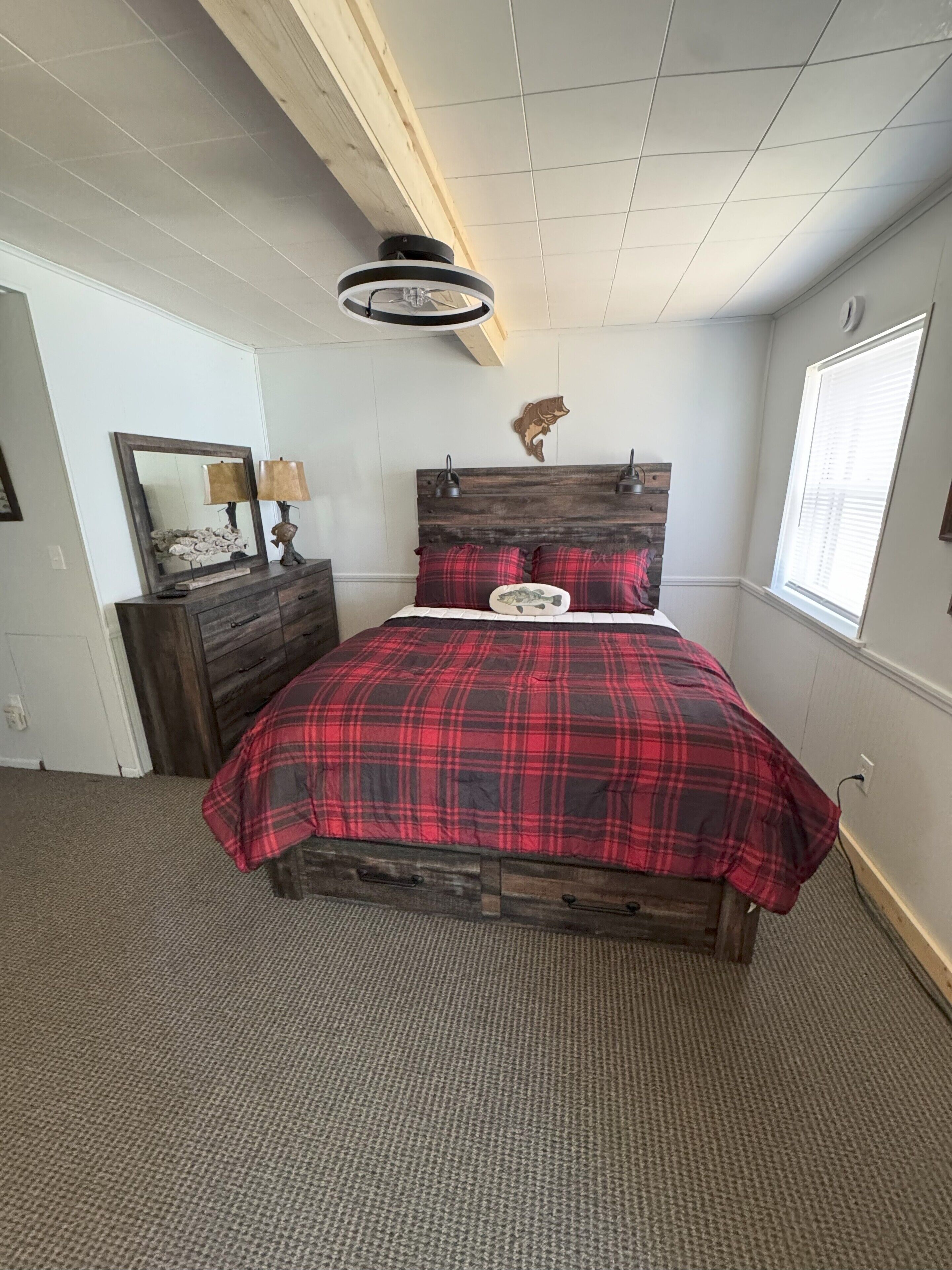 A bedroom with a wooden headboard, a red plaid bedspread, a wooden dresser, a mirror, and a wall-mounted cat head decoration.