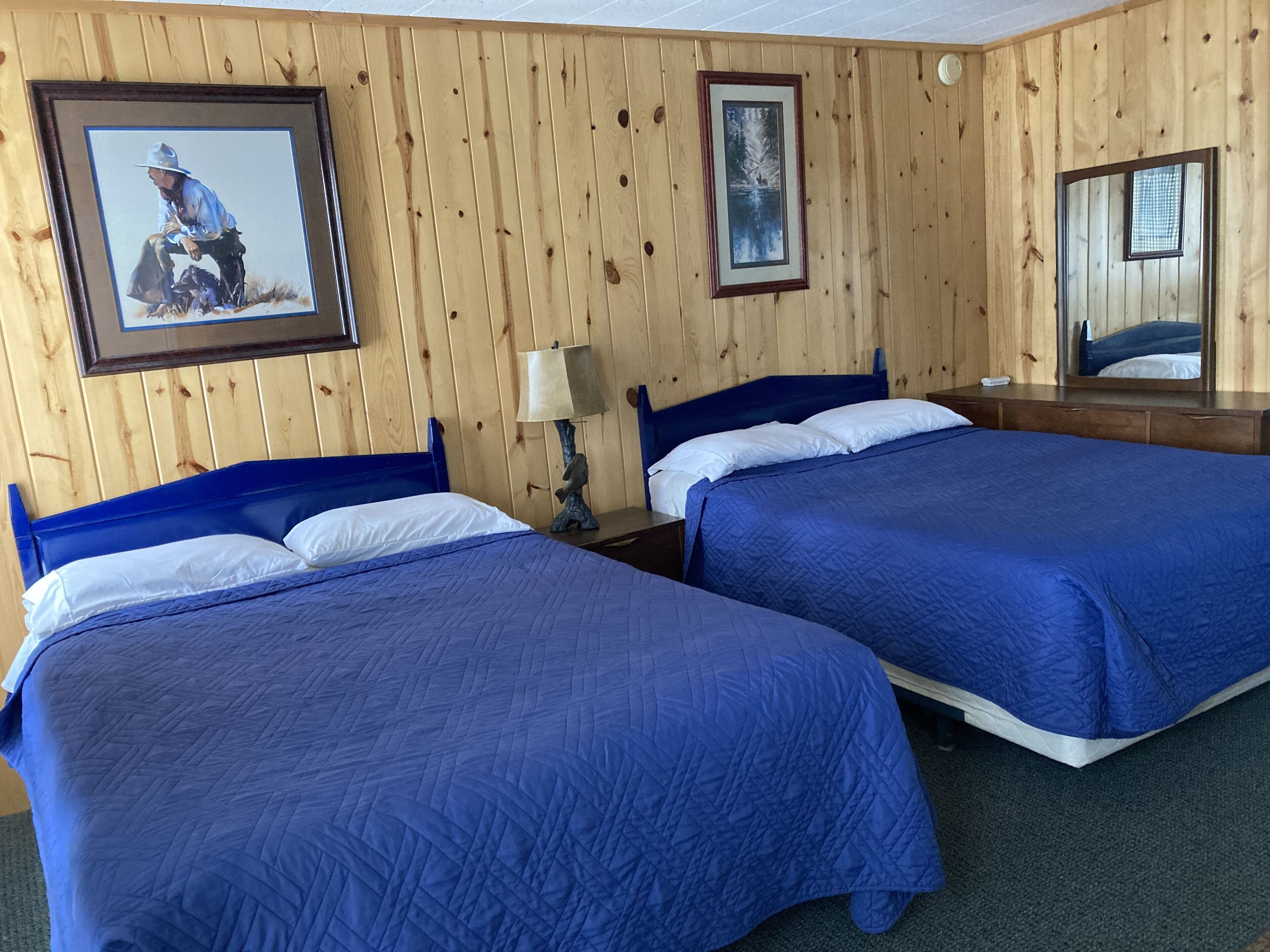 A room with two beds, wooden walls, a framed picture, a lamp, and a mirror.
