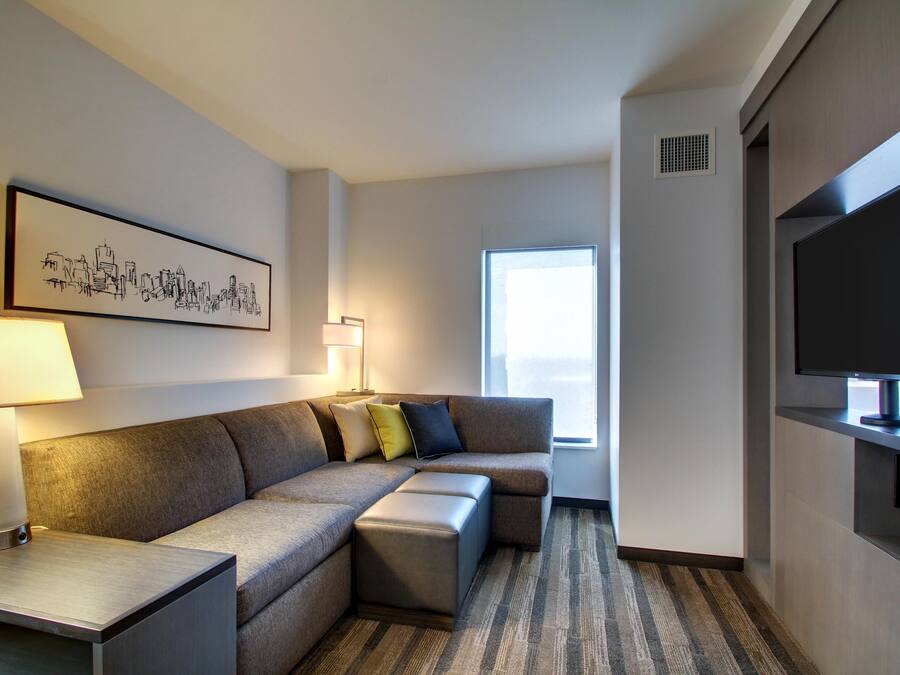 Ein modernes Hotelzimmer mit Ecksofa, Flachbildfernseher, Lampe und einem Stadtbild an der Wand.