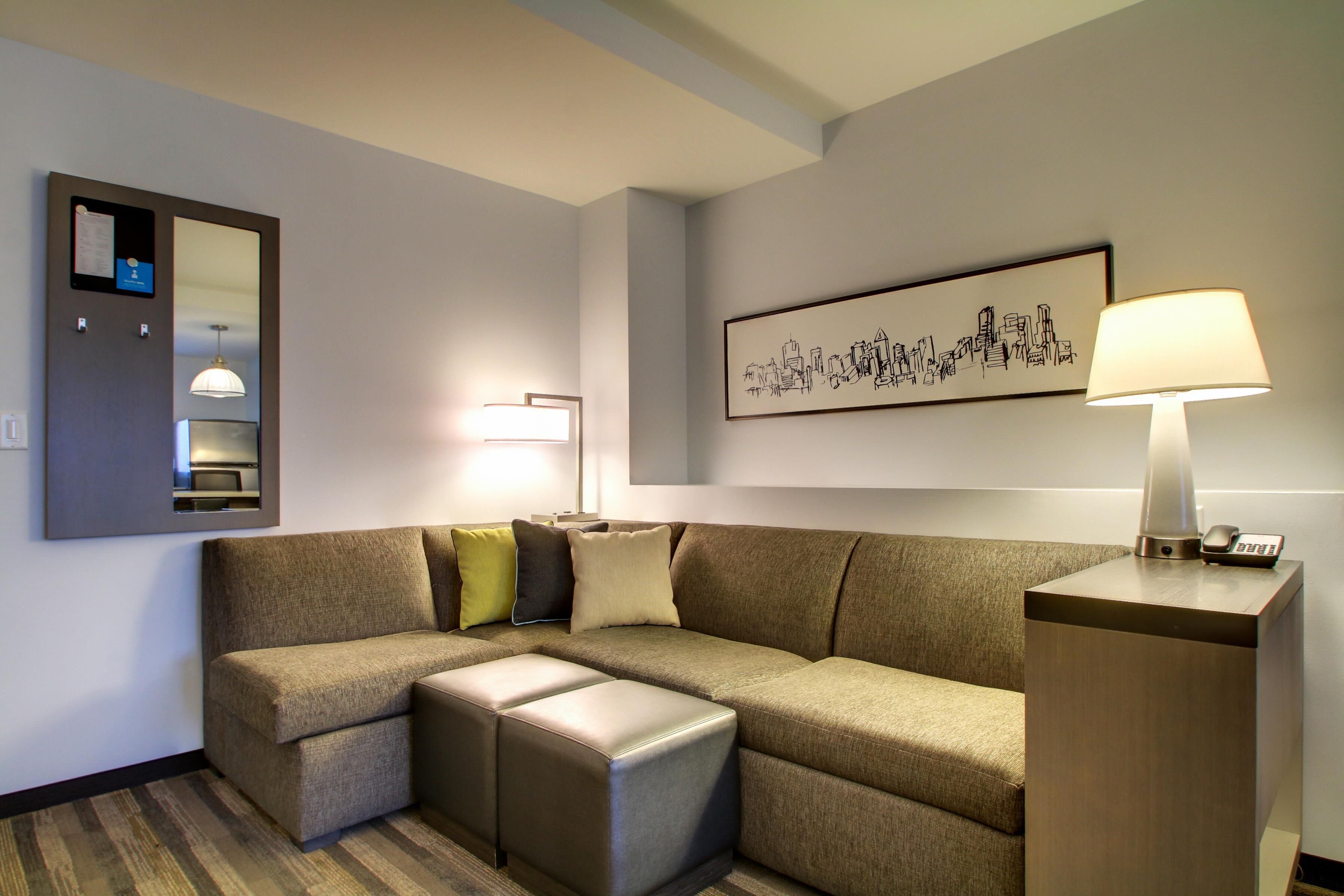 Une chambre d’hôtel moderne, dotée d’un canapé d’angle, d’une lampe sur pied, d’une œuvre d’art représentant un paysage urbain encadrée et d’une horloge numérique posée sur une table de chevet.