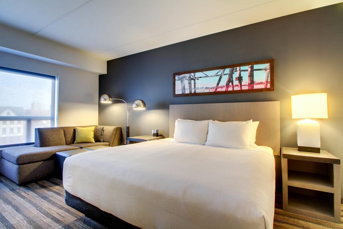 A hotel room with a large bed, a sofa, a desk, a lamp, and a painting on the wall.