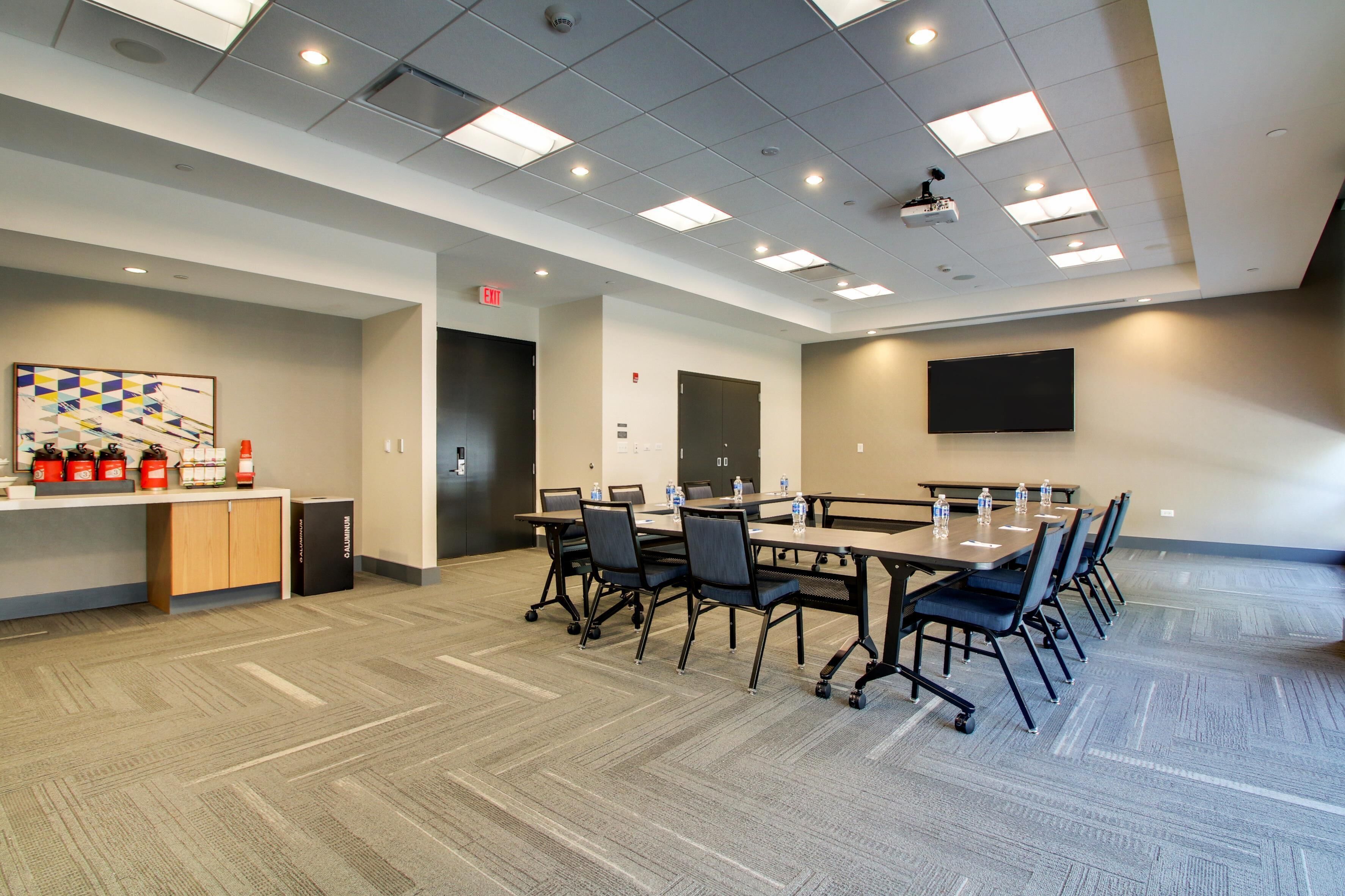A conference room with a long table, chairs, a flat-screen TV, and a small kitchenette area.
