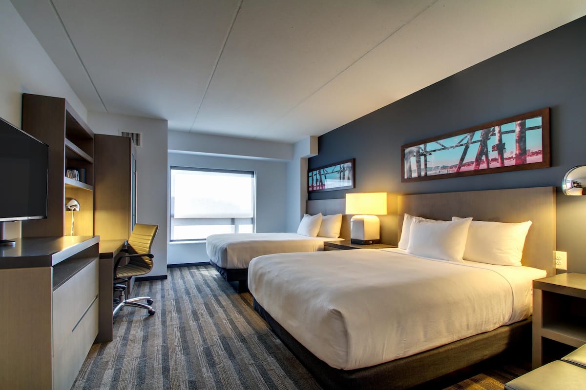 A hotel room with two beds, a desk, a chair, a TV, and artwork on the walls.