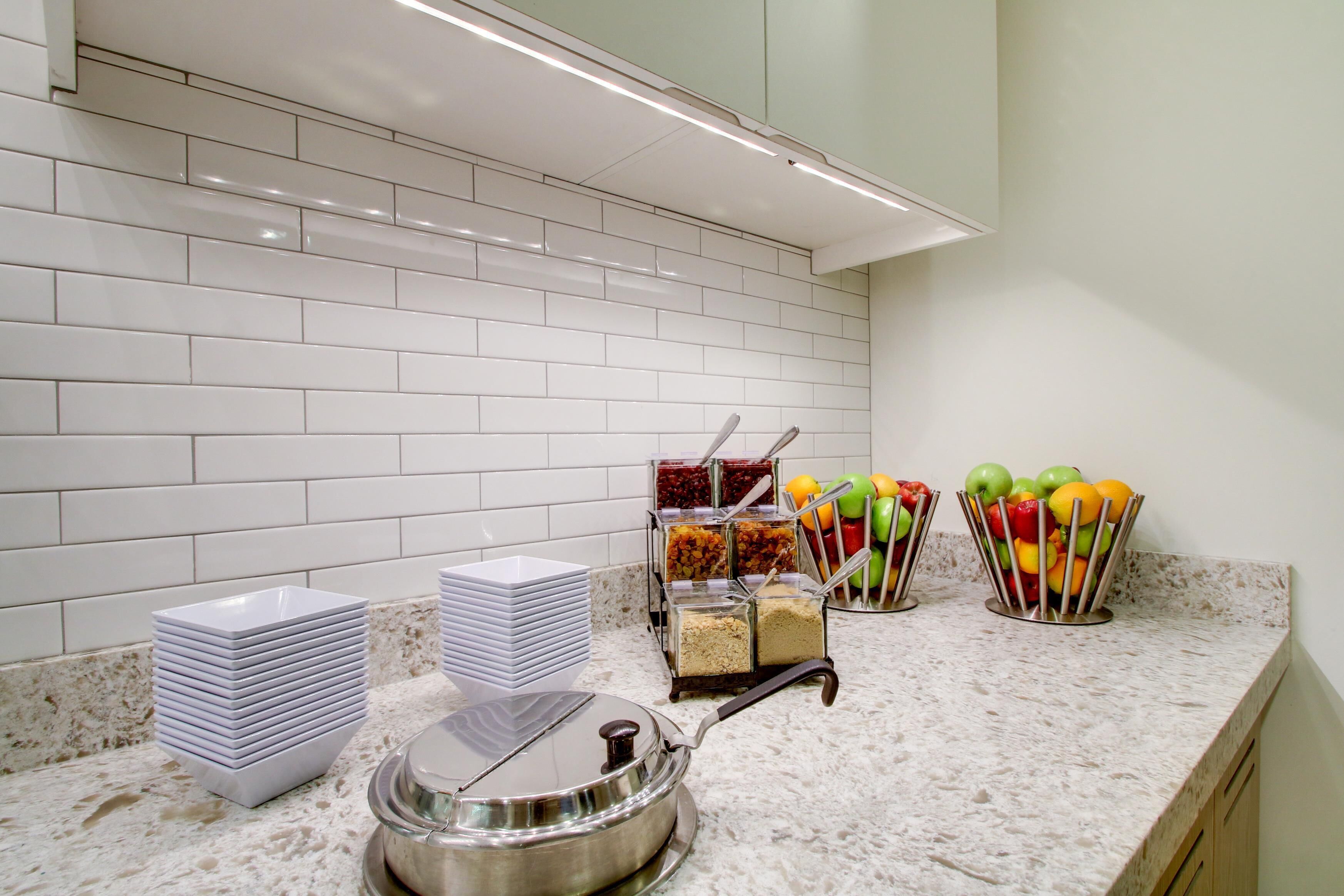 A kitchen area with white tiled backsplash, a granite countertop, stacked white plates, a stainless steel pot, and fruit containers.