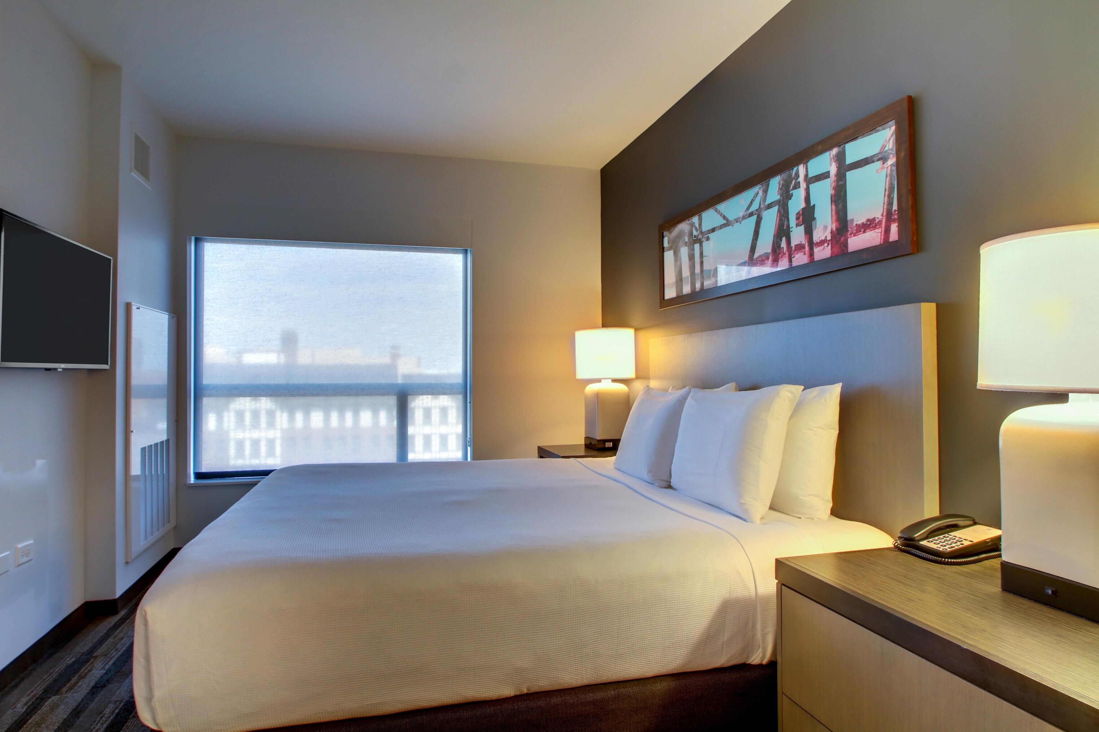 Habitación de hotel con una cama grande, televisión, ventana con vistas, un cuadro en la pared y una lámpara de noche.