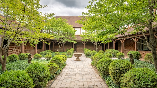 A courtyard with a brick pathway, surrounded by neatly trimmed hedges and trees, leading to a building with a tiled roof and brick walls.