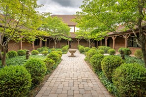 A courtyard with a brick pathway, surrounded by neatly trimmed hedges and trees, leading to a building with a tiled roof and brick walls.
