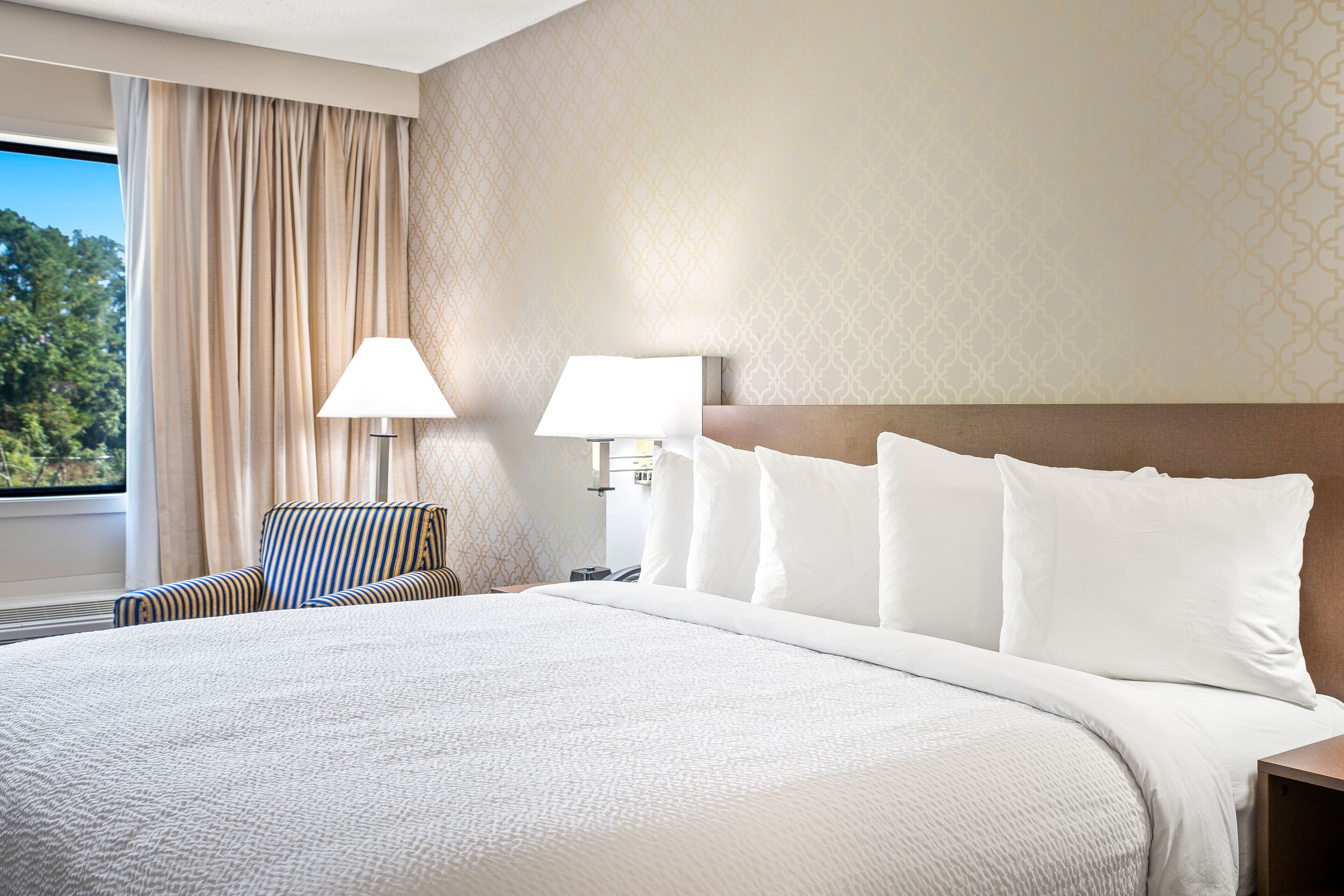 A hotel room with a large bed, two bedside lamps, a striped armchair, and a view of trees through a window with curtains.