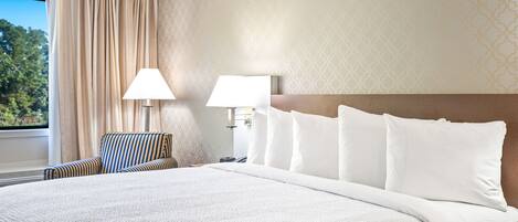 Habitación de hotel con una cama grande, dos lámparas de noche, un sillón a rayas y vistas a árboles a través de una ventana con cortinas.