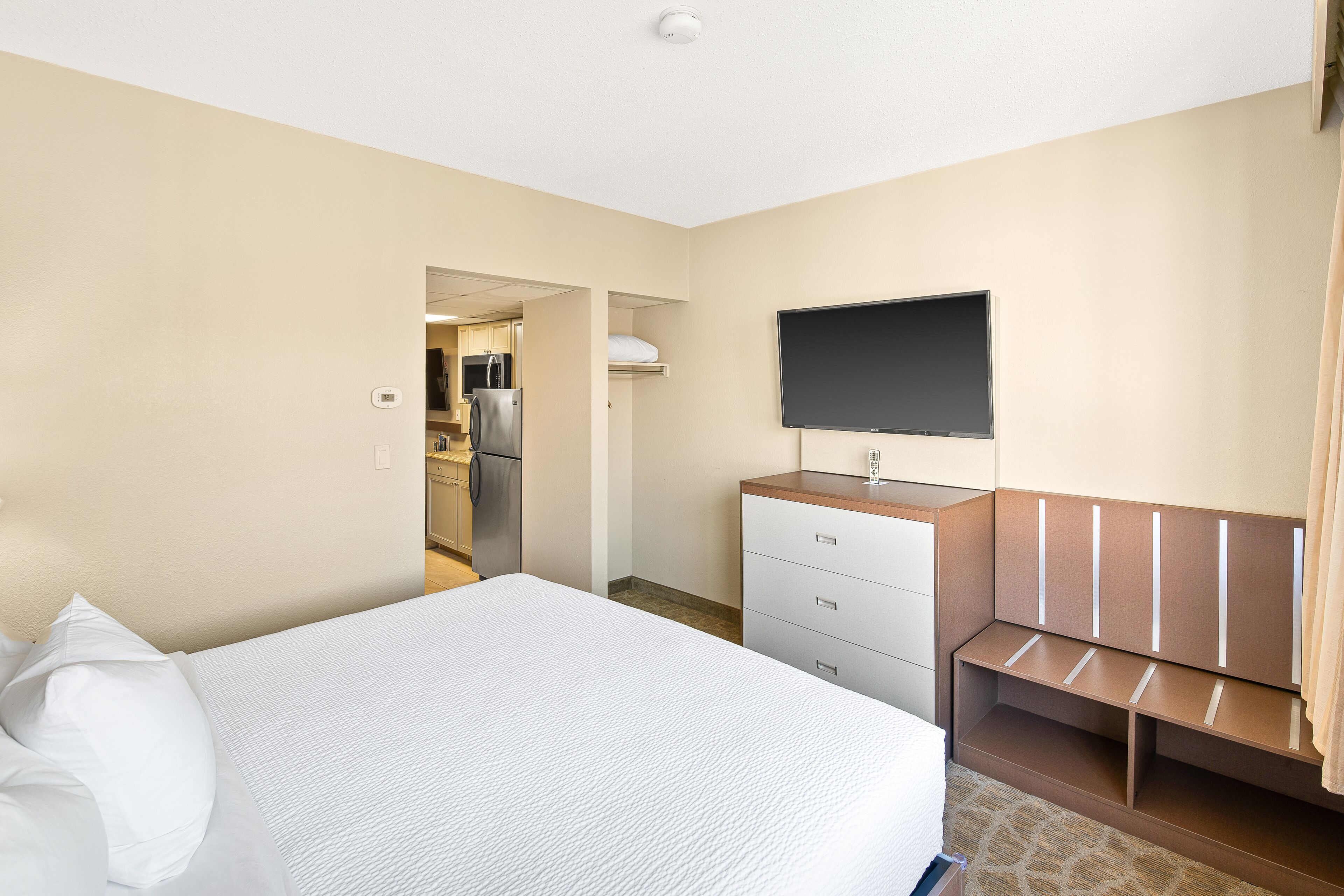 Habitación de hotel con cama, televisor montado en la pared, escritorio con cajones y una kitchenette al fondo.