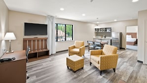 A modern hotel room with a flat-screen TV, a desk with a lamp, a seating area with two armchairs and a ottoman, a kitchenette, and a large window with a view of greenery.