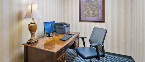 A wooden desk with a lamp, a computer, and a printer. There is a framed artwork on the wall and a checkered carpet.