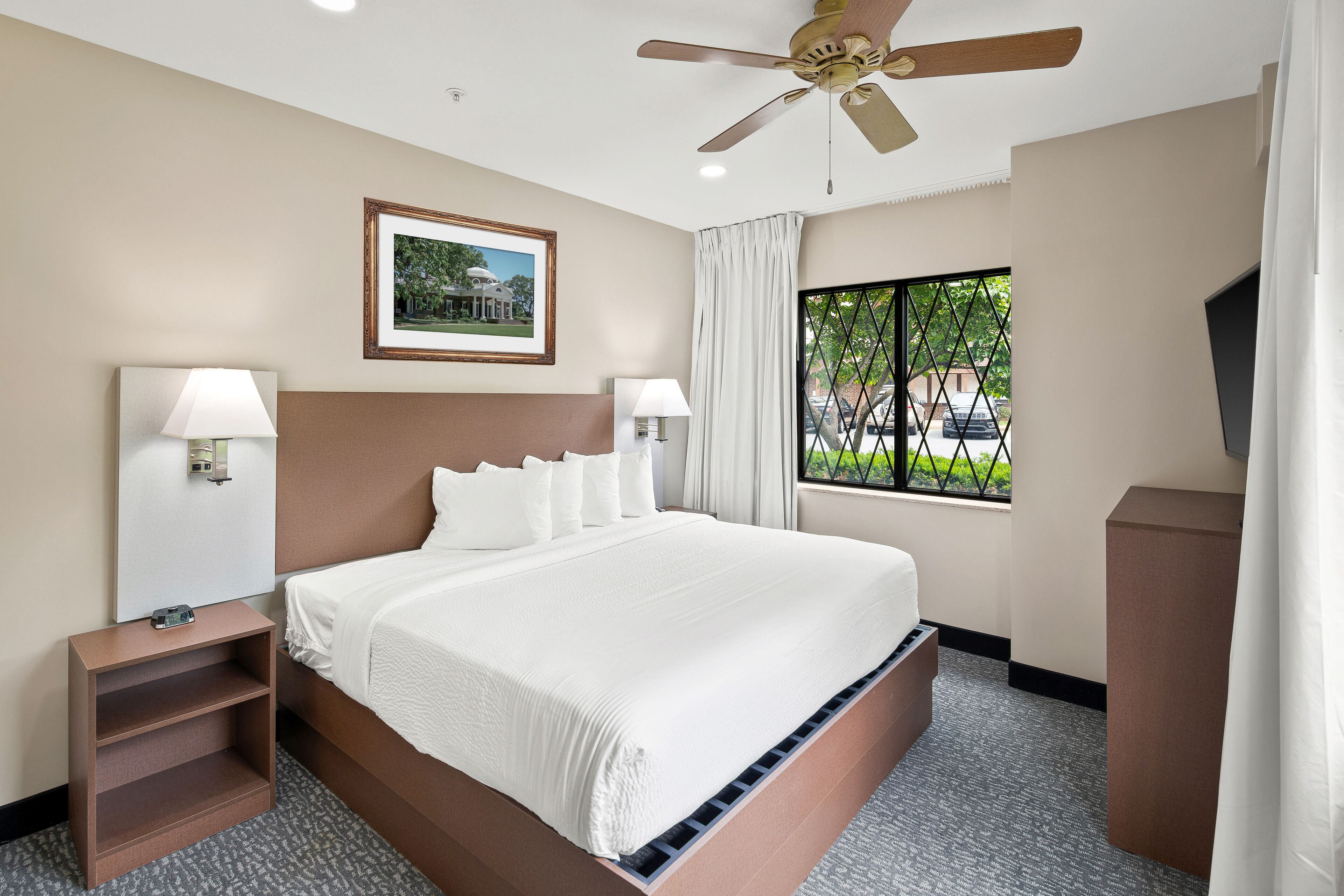 A hotel room with a large bed, two bedside lamps, a ceiling fan, a framed picture on the wall, a window with curtains, and a small wooden nightstand.