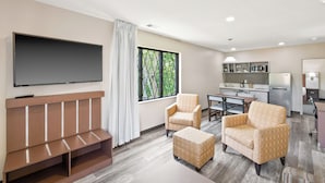 A modern living room with a flat-screen TV, two armchairs, a sofa, and a dining area with a kitchen in the background.