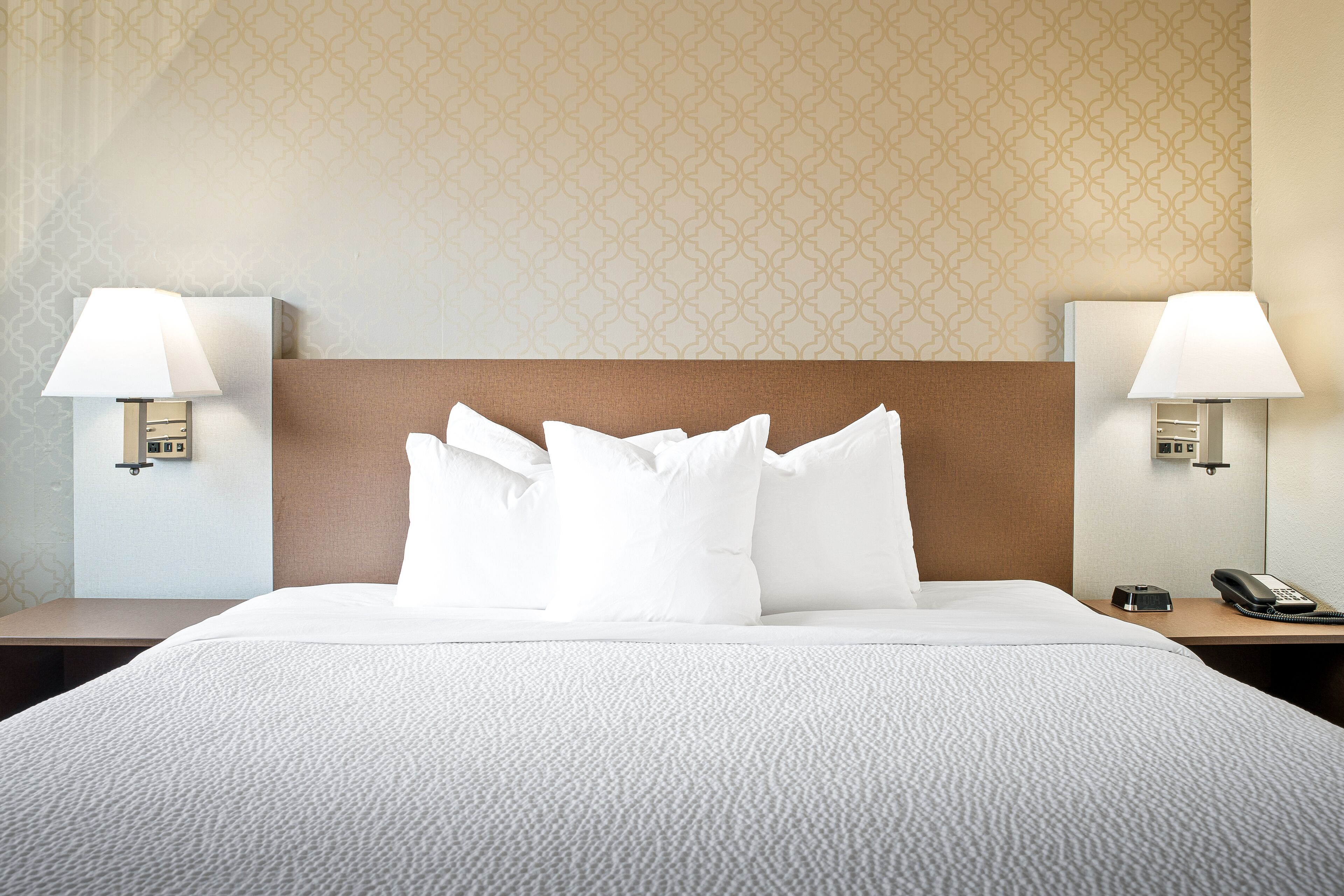 A neatly made bed with white linens and pillows, two bedside lamps, a telephone, and a patterned wall in the background.