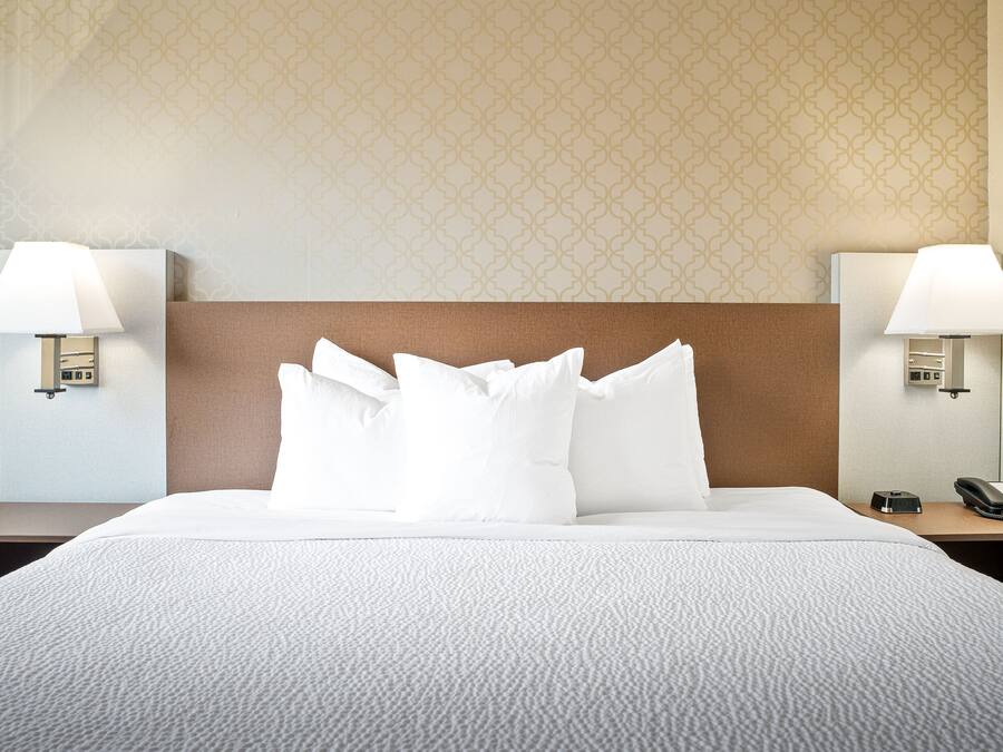 Una cama bien tendida con sábanas y almohadas blancas, dos lámparas de noche, un teléfono y un papel tapiz estampado al fondo.