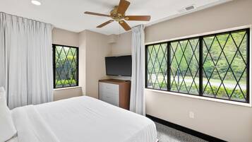 Habitación de hotel con cama, televisión, ventilador de techo y grandes ventanales con vistas a zonas verdes.