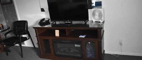 A room with a television on a wooden stand, a white fan, and a black chair.