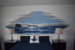 Un dormitorio con una cama, dos mesitas de noche con lámparas, una mesita auxiliar y un mural marino.