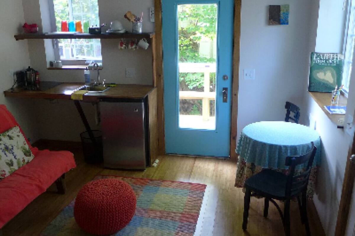 Eine kompakte Küche mit Kühlschrank, Spüle und einem Fenster über der Arbeitsfläche. Ein Esstisch mit einem Stuhl, ein roter Hocker und ein kleiner Teppich sind ebenfalls vorhanden.